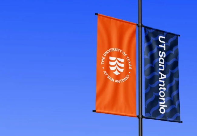 Two flags on a pole, a orange and blue, with the University of Texas at San Antonio logo on them